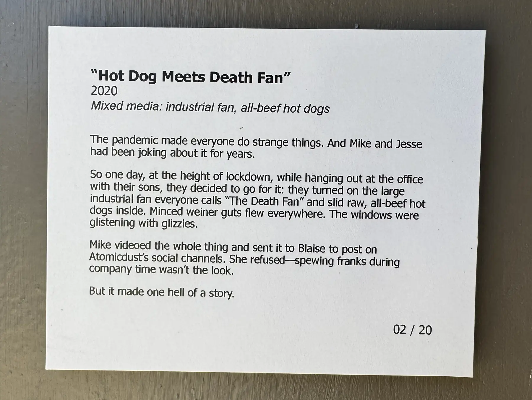 Printed museum-style exhibit placard titled “Hot Dog Meets Death Fan” resting on a metal surface